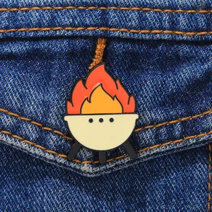 Detail of a cartoon BBQ grill enamel pin with orange flames on dark blue denim fabric