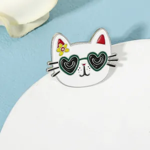 Close-up of a white enamel cat brooch wearing green heart-shaped sunglasses with a yellow flower