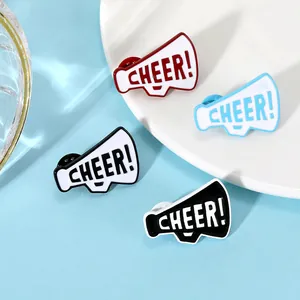 Dettaglio macro di spille a megafono in smalto con scritta 'CHEER!' su piatto bianco