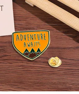 Gros plan détaillé du pin's émaillé Adventure Awaits avec montagnes sur bois sombre
