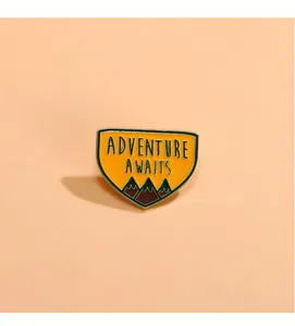 Gros plan sur un pin's émaillé jaune avec texte Adventure Awaits et motif de montagne