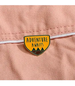 Gros plan d'un pin's émaillé jaune Adventure Awaits sur un tissu en denim rose pâle