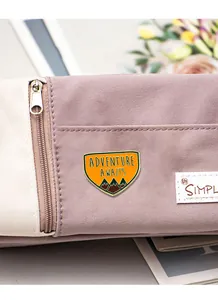 Détail d'une broche en émail orange avec texte Adventure Awaits sur un sac en toile violet