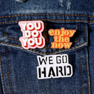 Dettaglio di tre spille smaltate su denim con citazioni YOU DO YOU e ENJOY THE NOW