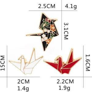 Three origami crane enamel pins in black floral, white, and red with gold metal edges