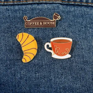 Detaillierte Emaille-Anstecknadeln mit Kaffeetasse und Croissant auf dunklem Denim