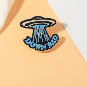 Detail showing a UFO enamel pin with blue drip oil finish and DOWN BAD typography text