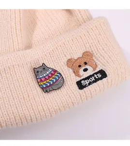 Detail of a cat enamel pin and bear embroidery on a cream knitted beanie cap