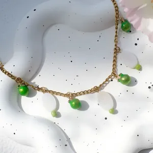 Detail van een gouden ketting met groene appelhanger en een witte parelmoeren bedel