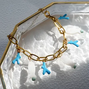 Detail van gouden schakelarmband met blauwe koraal en witte vis hangers op glazen dienblad