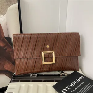 Pochette enveloppe marron plissée avec grande boucle dorée posée sur une valise blanche