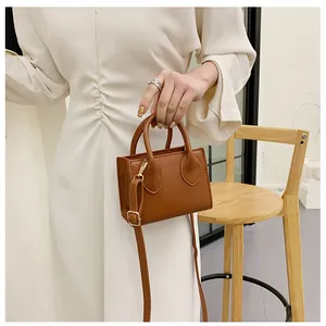 Brown mini square handbag with dual top handles and long crossbody strap shown on a chair