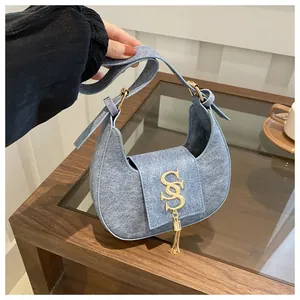 Détail du sac croissant en denim avec logo SS doré et pendentif à franges sur table