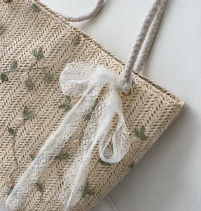 Detail of straw tote bag featuring green leaf embroidery and a large white lace bow
