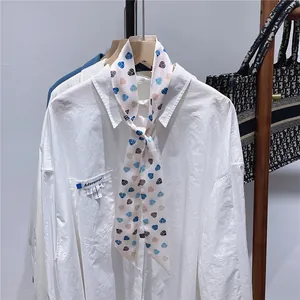 Detail einer weißen Bluse mit Herzmuster-Bandschal auf einem Holzbügel präsentiert
