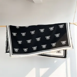 Detail of a black knit shawl featuring white bee patterns and delicate fringe hanging on wood