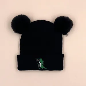 Close-up of black knit baby beanie with double faux fur pom-poms and green crocodile embroidery