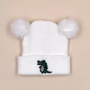 Detail of white double pom-pom knit baby hat featuring a green crocodile embroidery