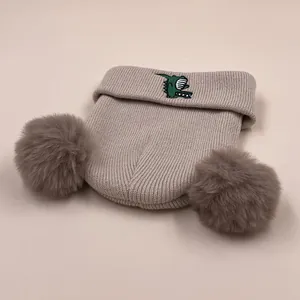 Grey knit winter hat for children with fluffy pom-poms and cute dinosaur embroidery