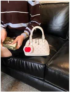 Detailansicht einer weißen gepunkteten Muscheltasche mit rotem Herzanhänger auf Ledersofa