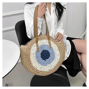 Detail of round straw tote bag with blue evil eye pattern and brown leather straps