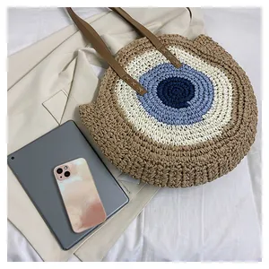 Detail of round woven straw tote featuring evil eye pattern and leather shoulder straps
