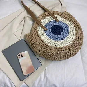 Round straw tote bag with evil eye pattern and brown leather straps for beach vacation