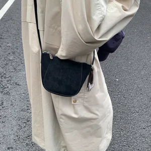 Detail einer schwarzen Halbmond-Umhängetasche aus Leder mit Quaste auf einem beigen Trenchcoat