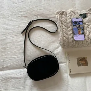 Flat lay of black oval suede crossbody bag with adjustable strap on a white fabric background