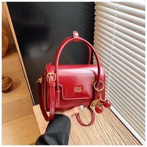 Detail of red leather crossbody bag with cherry charm and elegant metal logo on wooden desk