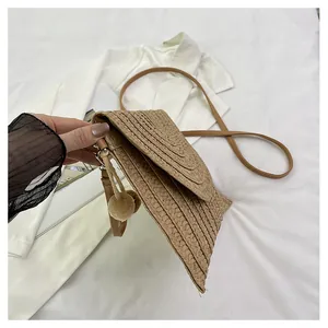 Detail of woven straw envelope crossbody bag featuring decorative beige pom-poms and brown strap