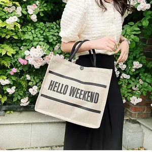 Beige canvas tote bag with black braided handles featuring Hello Weekend print outdoors