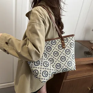 Large beige tote bag with M logo pattern and brown straps, worn with a trench coat