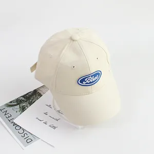 Top-down view of cream cotton baseball cap with blue embroidered letter patch and air holes