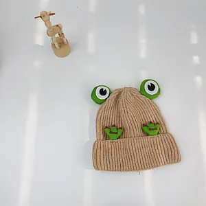 Detail einer beige Strickmütze für Babys mit 3D-Froschaugen und Good-Stickerei auf weiß