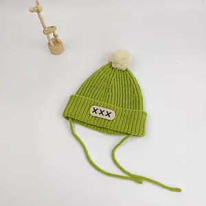 Detail of green knit baby hat with white pom-pom, letter patch and side straps