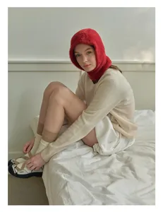 Detail of red plush balaclava hat paired with beige knit sweater in a cozy indoor setting