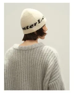 Nahaufnahme der cremefarbenen Strickmütze mit Jacquard-Buchstaben und grauem Pullover