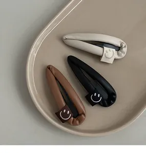 Three-piece PU leather water drop hair clips with smiley face tags on a ceramic tray