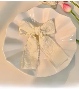 Detail of cream bow hair clip with pearl trim and pleated texture on a white shell plate