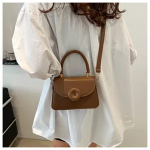 Brown leather crossbody bag with top handle shown on a white shirt in a modern room