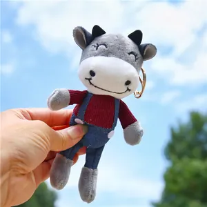 Cute plush cow keychain toy in red corduroy top and blue overalls on sunny background
