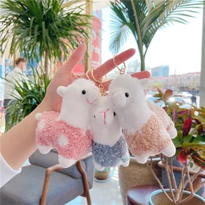 Three cute alpaca plush keychains with gold metal clips held by hand in a sunny garden