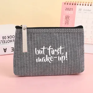 Detail of grey striped canvas makeup bag featuring 'but first, make-up!' text and leather strap