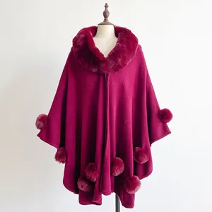 Detail of burgundy knit poncho featuring a plush faux fur collar and decorative pom-poms