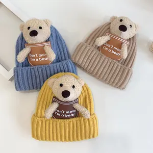 Three knitted baby hats in blue, khaki, and yellow with plush bear detail and leather tags