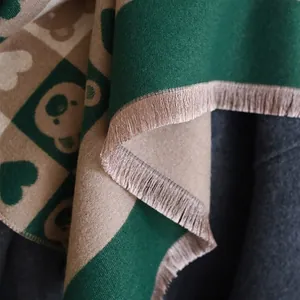 Détail d'une écharpe à carreaux vert et beige avec motif ours et pompons à franges