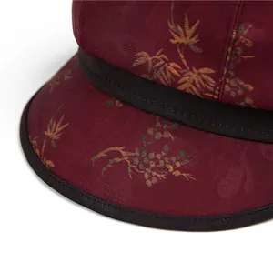 Detail eines weinroten Stoffes mit goldenem Blattmuster und schwarzer Paspelierung am Damenhut