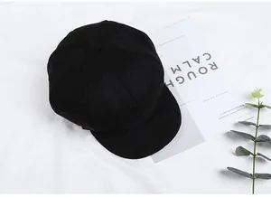 Top view of a black octagonal newsboy hat on a white textured background with green leaves