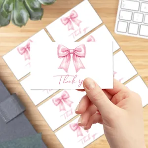 Hand holding a white thank you card with a pink watercolor bow design on a desk